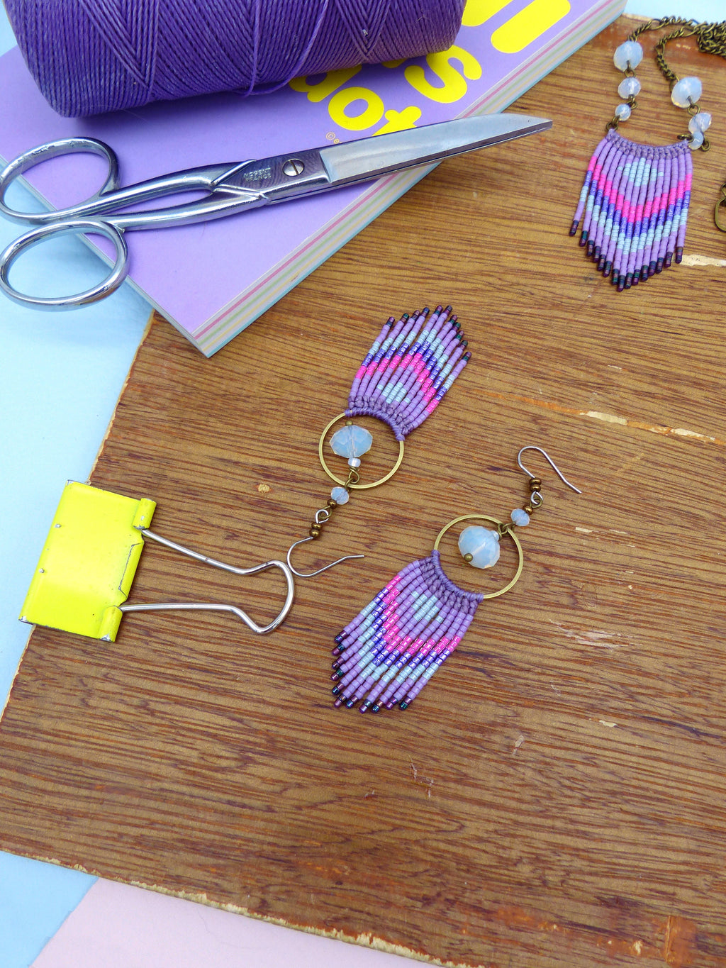 Peut inclure: Une paire de boucles d'oreilles en perles violettes, roses et bleues avec une pierre blanche et un anneau doré. Les boucles d'oreilles sont sur une surface en bois.