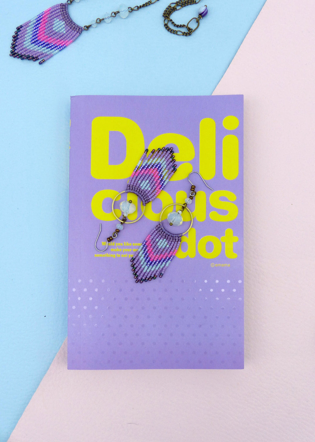 Peut inclure: Une paire de boucles d'oreilles en perles violettes et bleues avec un anneau et un pendentif en forme de larme. Les boucles d'oreilles sont sur un livre violet avec le texte "Delicious dot" en jaune.