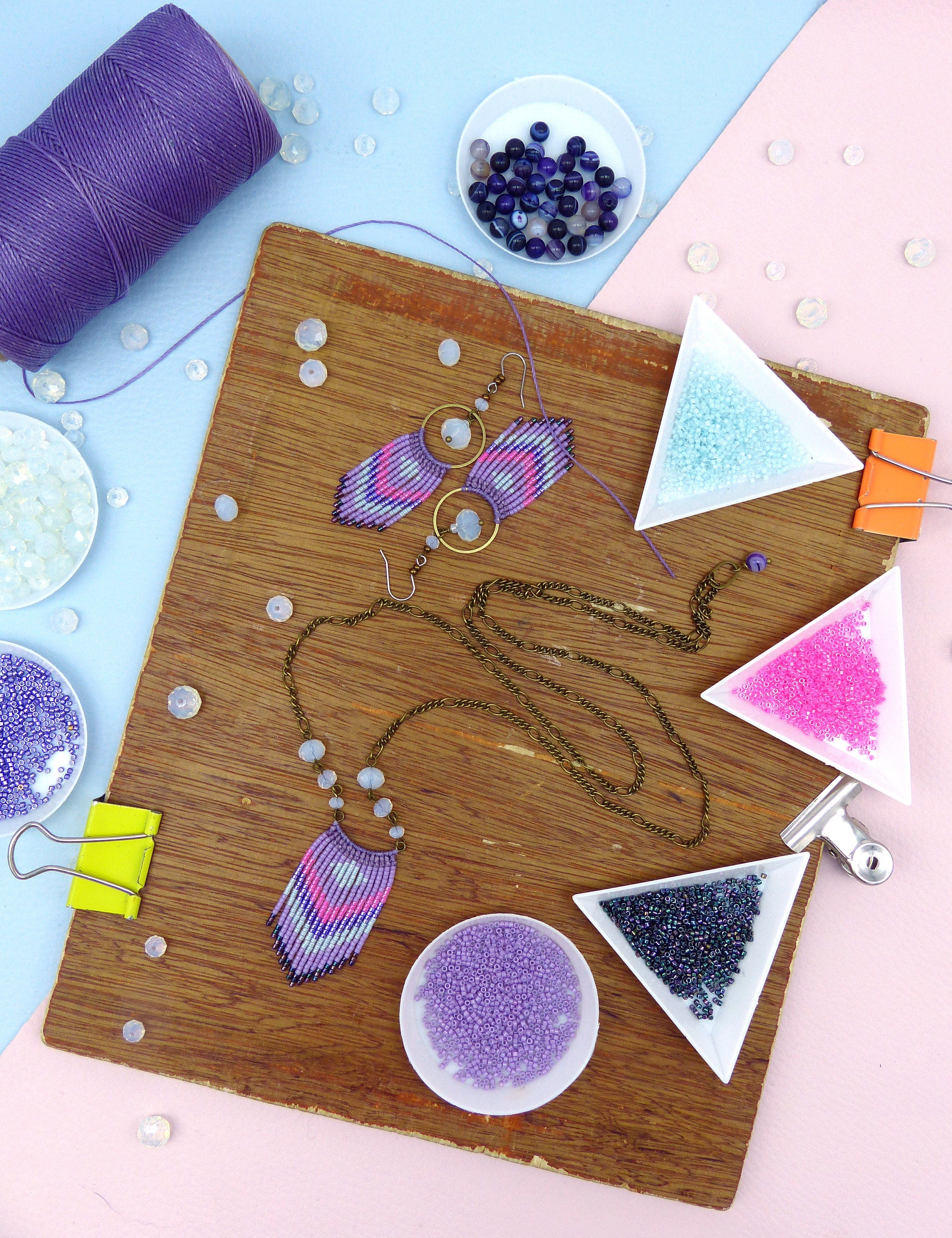 Peut inclure: Un plateau en bois avec divers perles et fournitures de bijouterie. Il y a de petits bols de perles en violet, rose, bleu et noir. Il y a aussi des boucles d'oreilles et un collier faits de perles dans des tons de violet, rose et bleu.