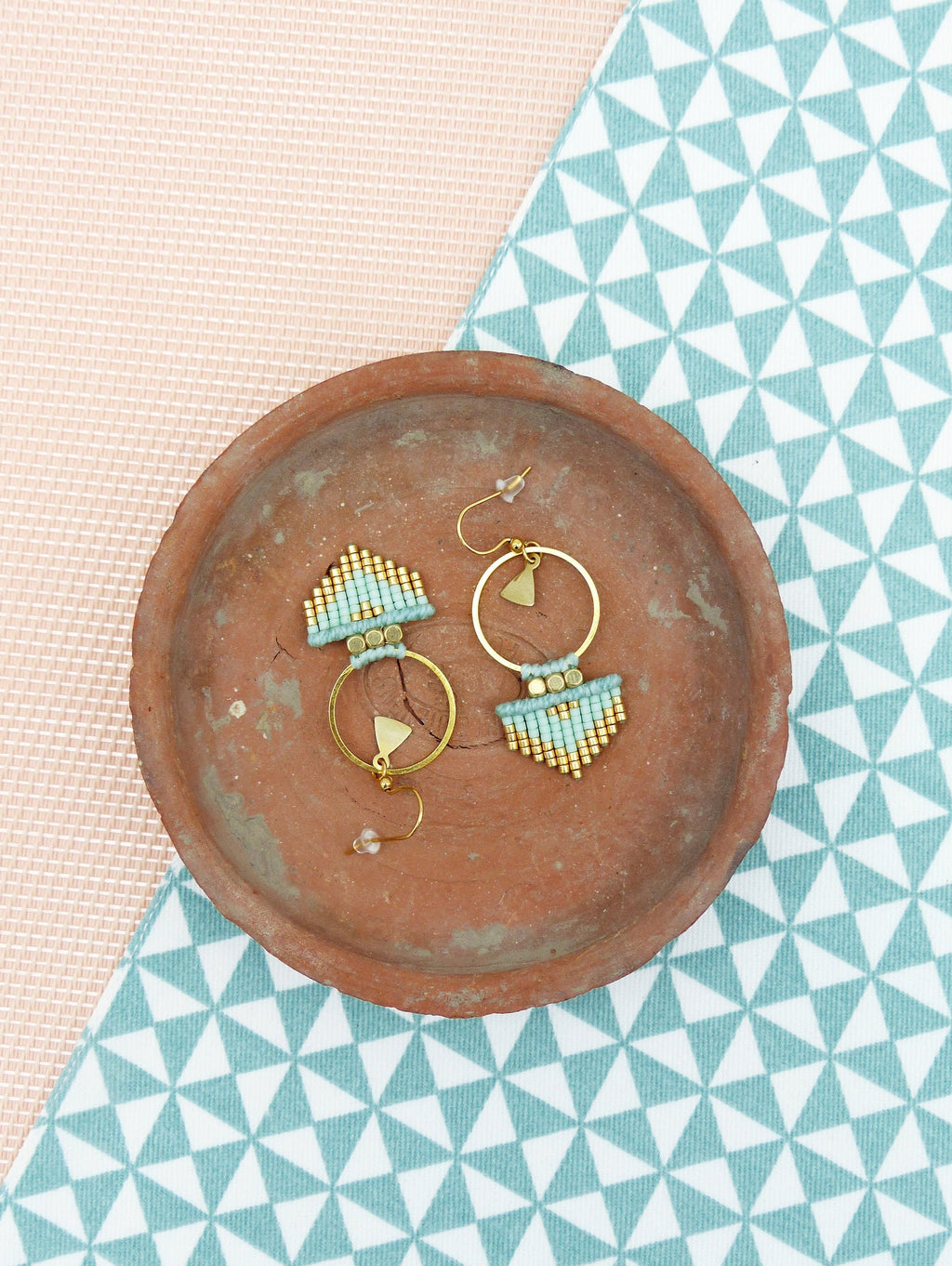 Peut inclure: Une paire de boucles d'oreilles créoles dorées avec des franges de perles turquoise et dorées. Les boucles d'oreilles sont présentées sur un plat en terre cuite.