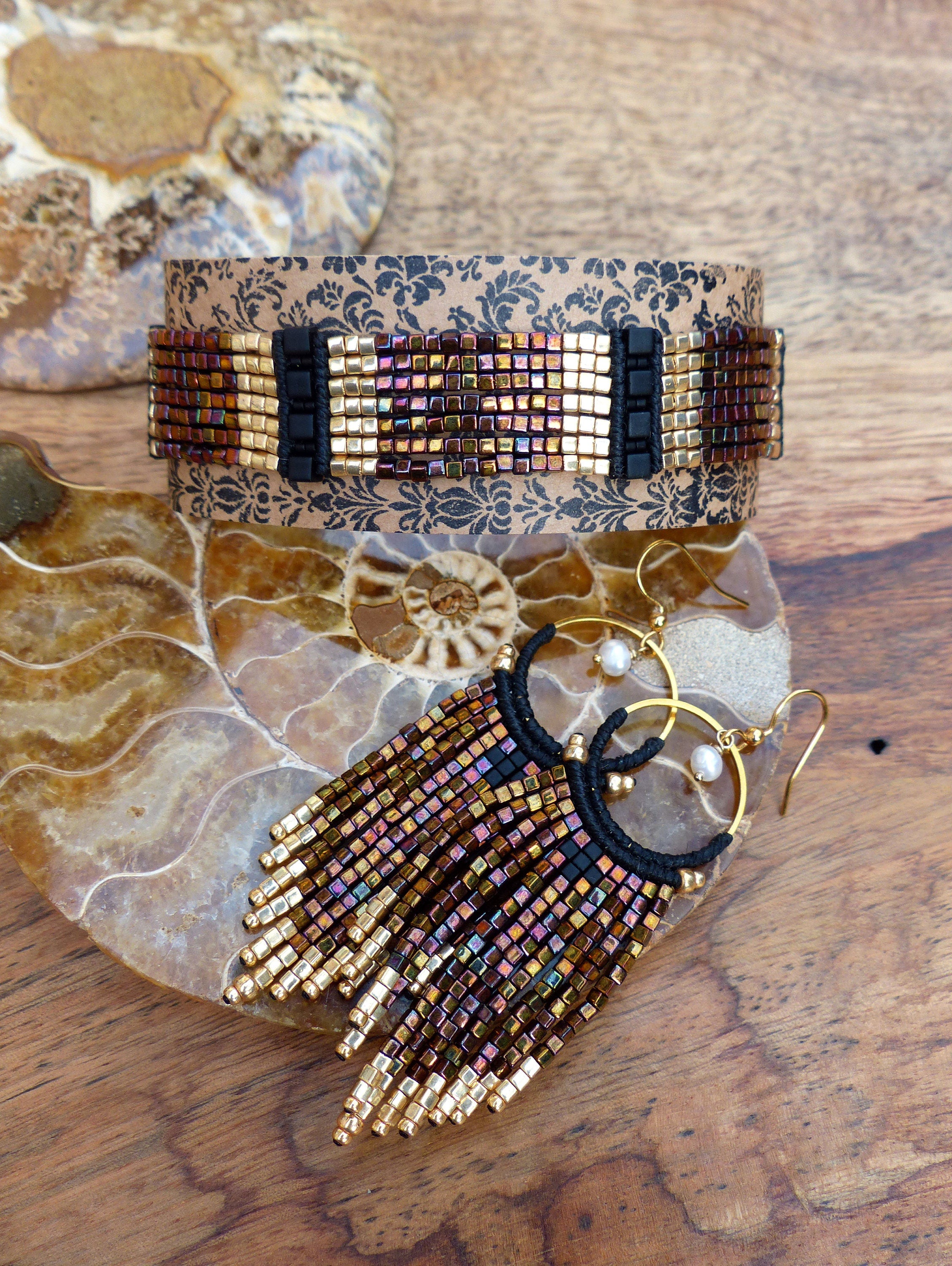 Peut inclure: Une paire de boucles d'oreilles créoles dorées avec une frange de perles noires et brunes. Les boucles d'oreilles sont présentées sur une surface à motifs brun et blanc.