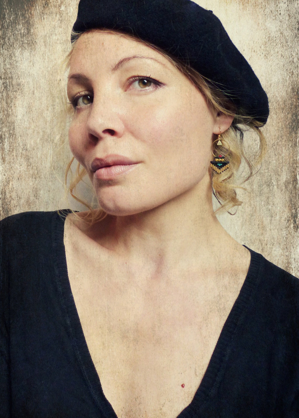 Peut inclure: Une femme aux cheveux blonds portant un béret noir et un chemisier noir à col en V. Elle porte des boucles d'oreilles en perles dorées et noires.