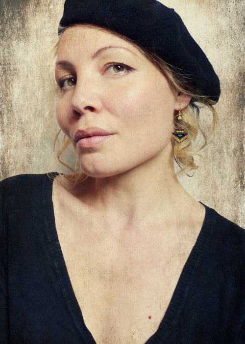 Peut inclure: Une femme aux cheveux blonds portant un béret noir et un chemisier noir à col en V. Elle porte des boucles d'oreilles en perles dorées et noires.