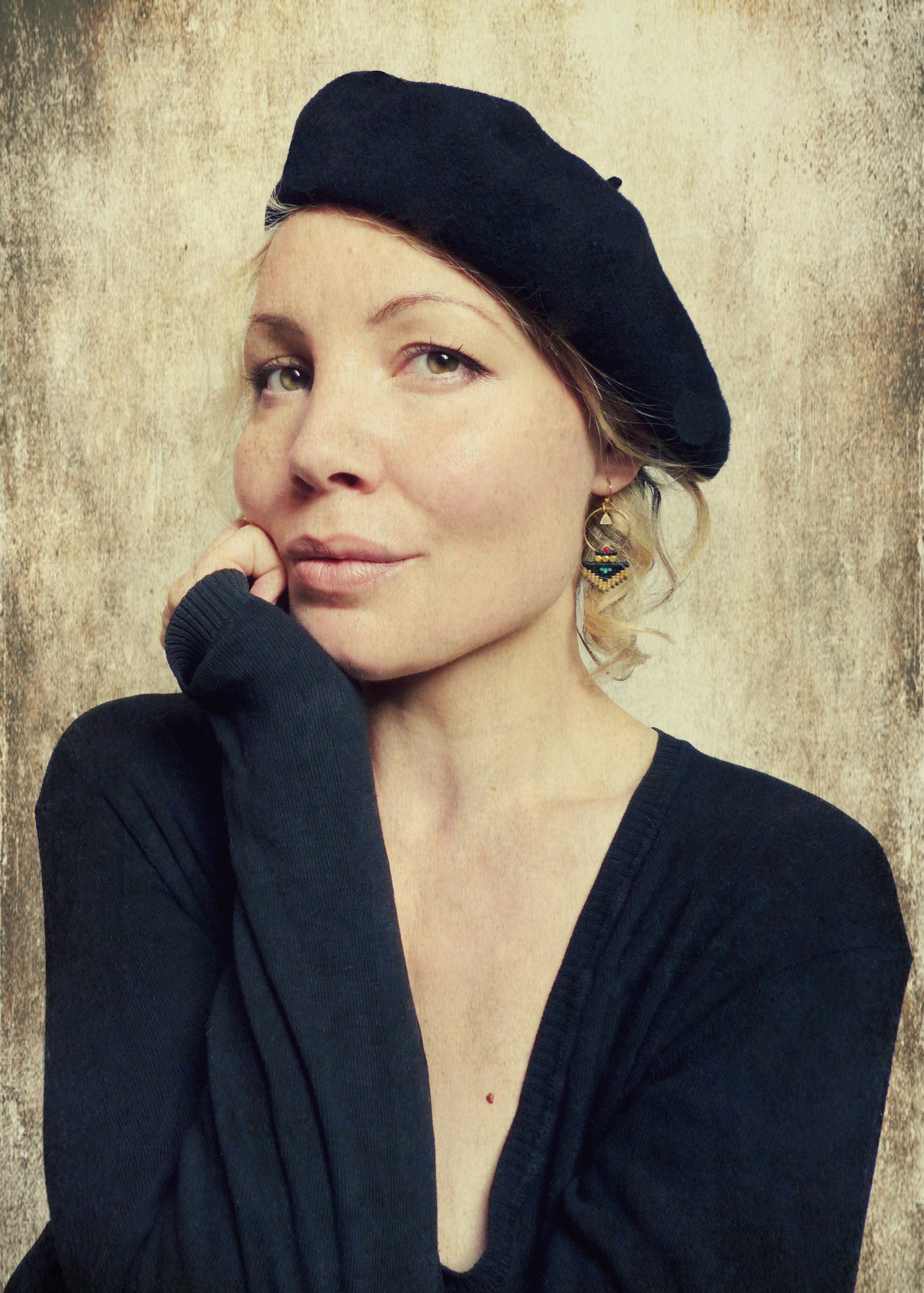 Peut inclure: Une femme aux cheveux bruns courts et ondulés porte un béret noir et un pull noir à col en V. Elle regarde la caméra et a une expression neutre. Elle porte de petites boucles d'oreilles dorées avec des accents turquoise et noirs.