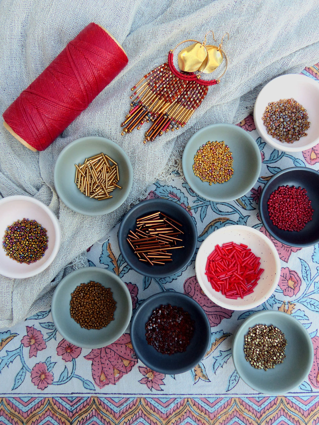 Peut inclure: Assortiment de perles, perles de rocaille et de composants métalliques dans de petits bols sur un tissu à motif floral. Les perles sont de différentes couleurs, notamment or, rouge, marron et irisé. Une bobine de fil rouge et une paire de boucles d'oreilles créoles dorées avec des franges de perles sont également visibles.