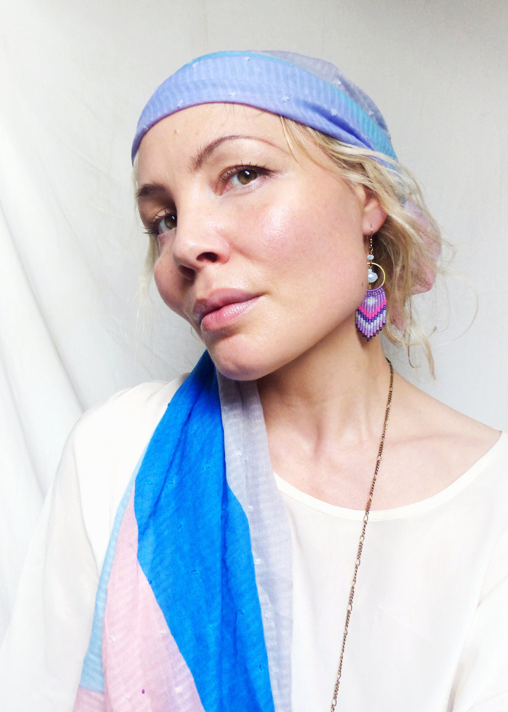 Peut inclure: Une femme portant une écharpe à motifs bleus et roses et un foulard bleu clair à pois blancs. Elle porte une chemise blanche et un collier de chaîne en or. Elle porte une paire de boucles d'oreilles en perles violettes et roses.