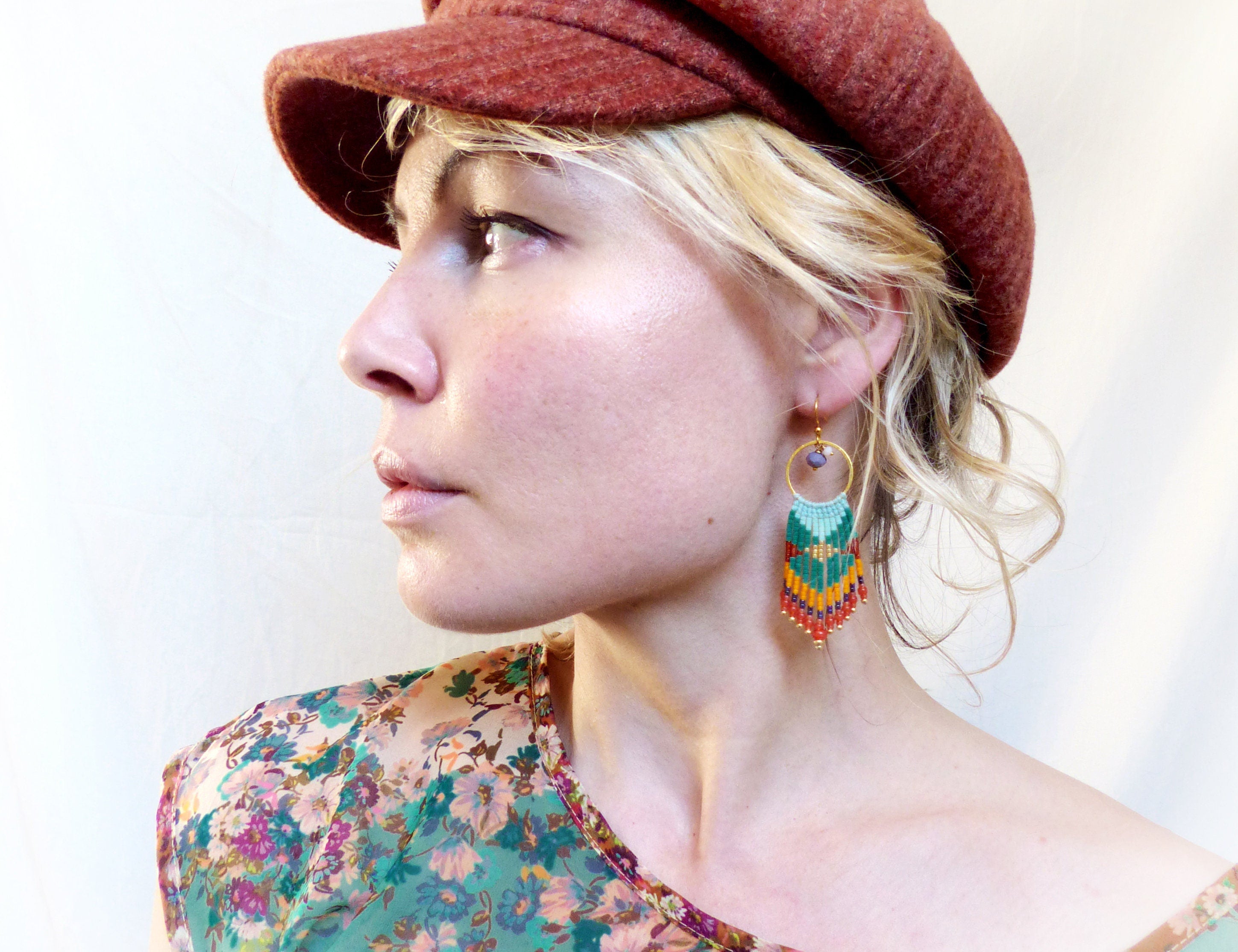 Peut inclure: Une femme portant une casquette rouge et une chemise à fleurs avec des boucles d'oreilles en perles colorées. Les boucles d'oreilles sont faites de petites perles dans des tons de vert, orange, rouge et jaune, avec un anneau en or en haut.