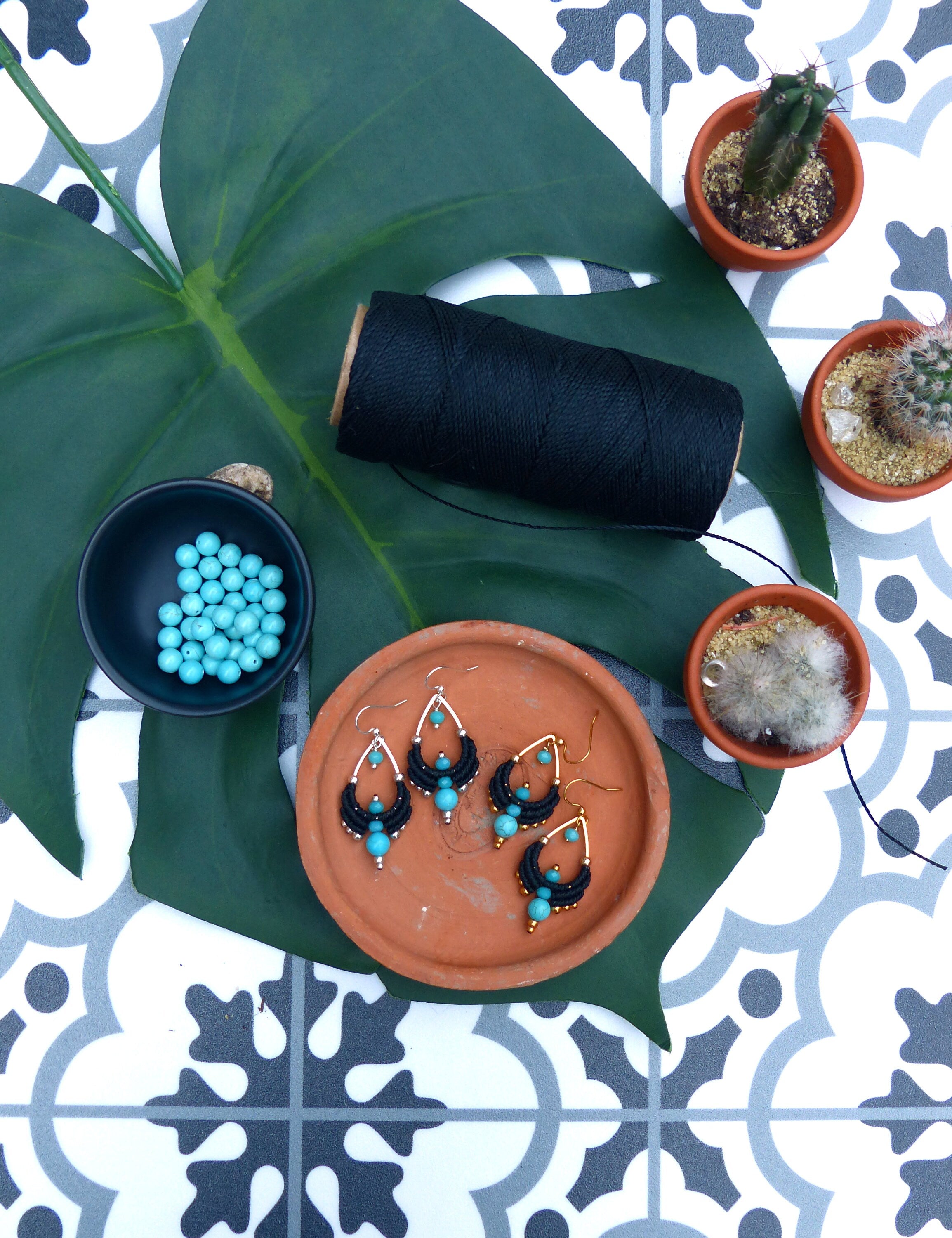 Peut inclure: Trois paires de boucles d'oreilles en turquoise et noires avec des accents dorés sur une assiette en terre cuite. Les boucles d'oreilles sont présentées sur une feuille verte avec une bobine de fil noir et de petites plantes succulentes en pot.