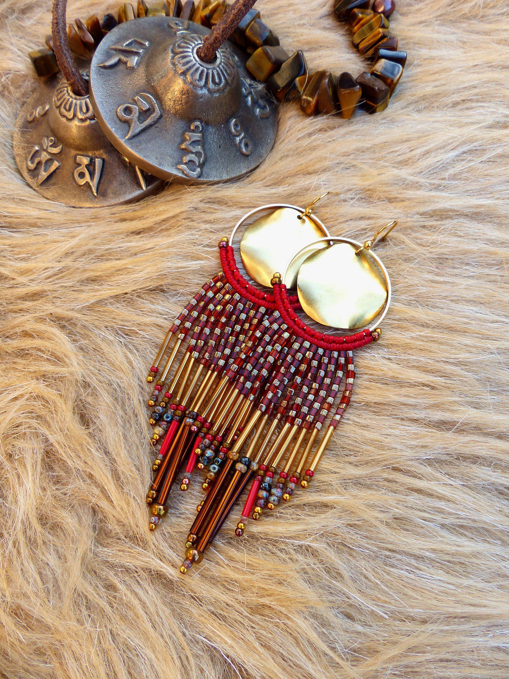 Peut inclure: Boucles d'oreilles créoles dorées avec une frange de perles rouges et dorées. Les boucles d'oreilles sont sur un fond de fourrure brune.