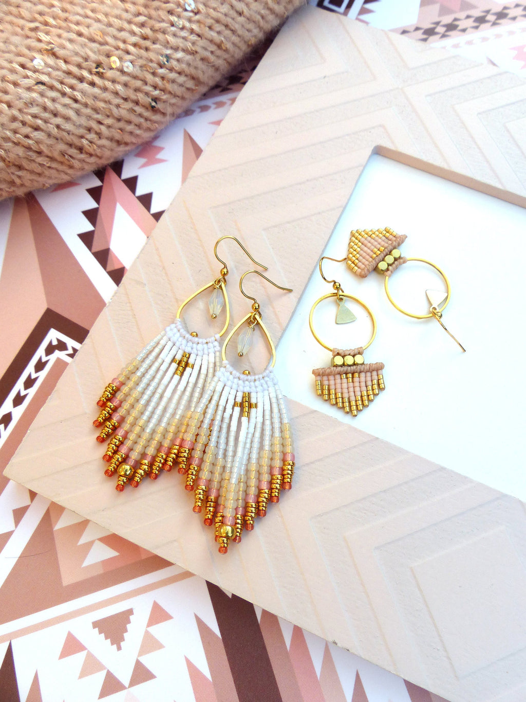 Peut inclure: Une paire de boucles d'oreilles en perles dorées et blanches avec un design en frange. Les boucles d'oreilles sont sur une surface blanche avec un motif géométrique.
