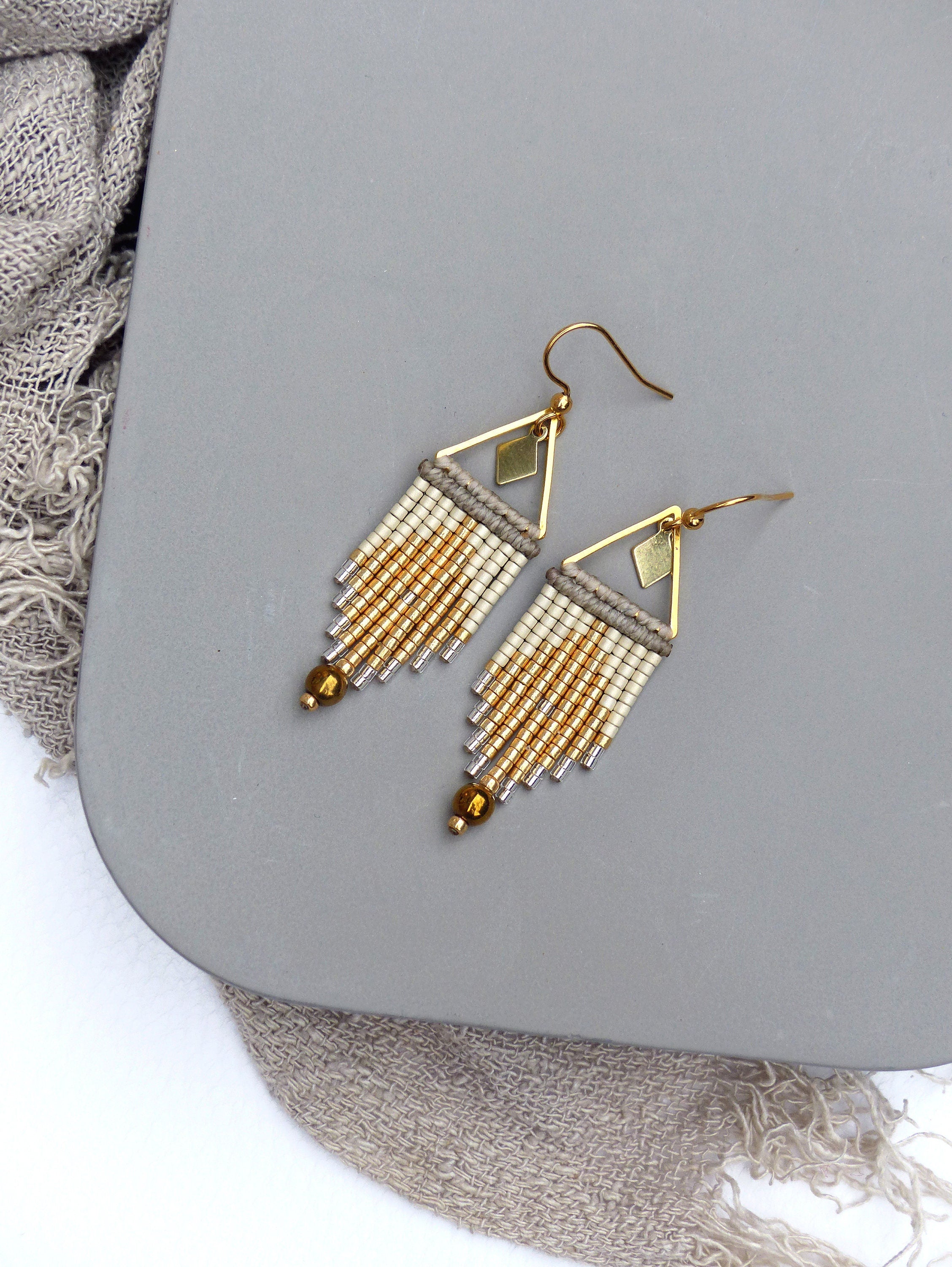 Peut inclure: Une paire de boucles d'oreilles en perles dorées et blanches avec un motif géométrique triangulaire. Les boucles d'oreilles sont faites de petites perles de rocaille et ont une frange pendante.