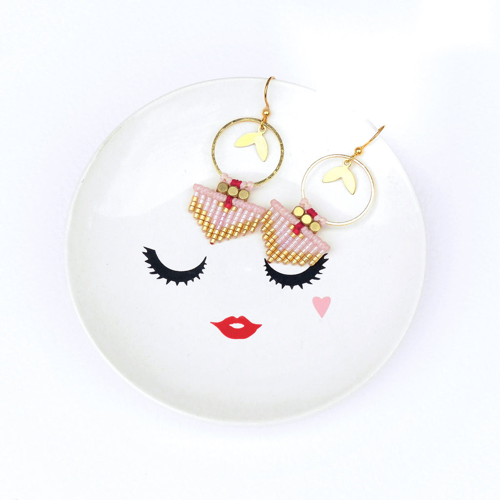 Peut inclure: Une paire de boucles d'oreilles créoles dorées avec des franges de perles roses et rouges. Les boucles d'oreilles sont sur une assiette blanche avec un motif de visage noir et blanc.