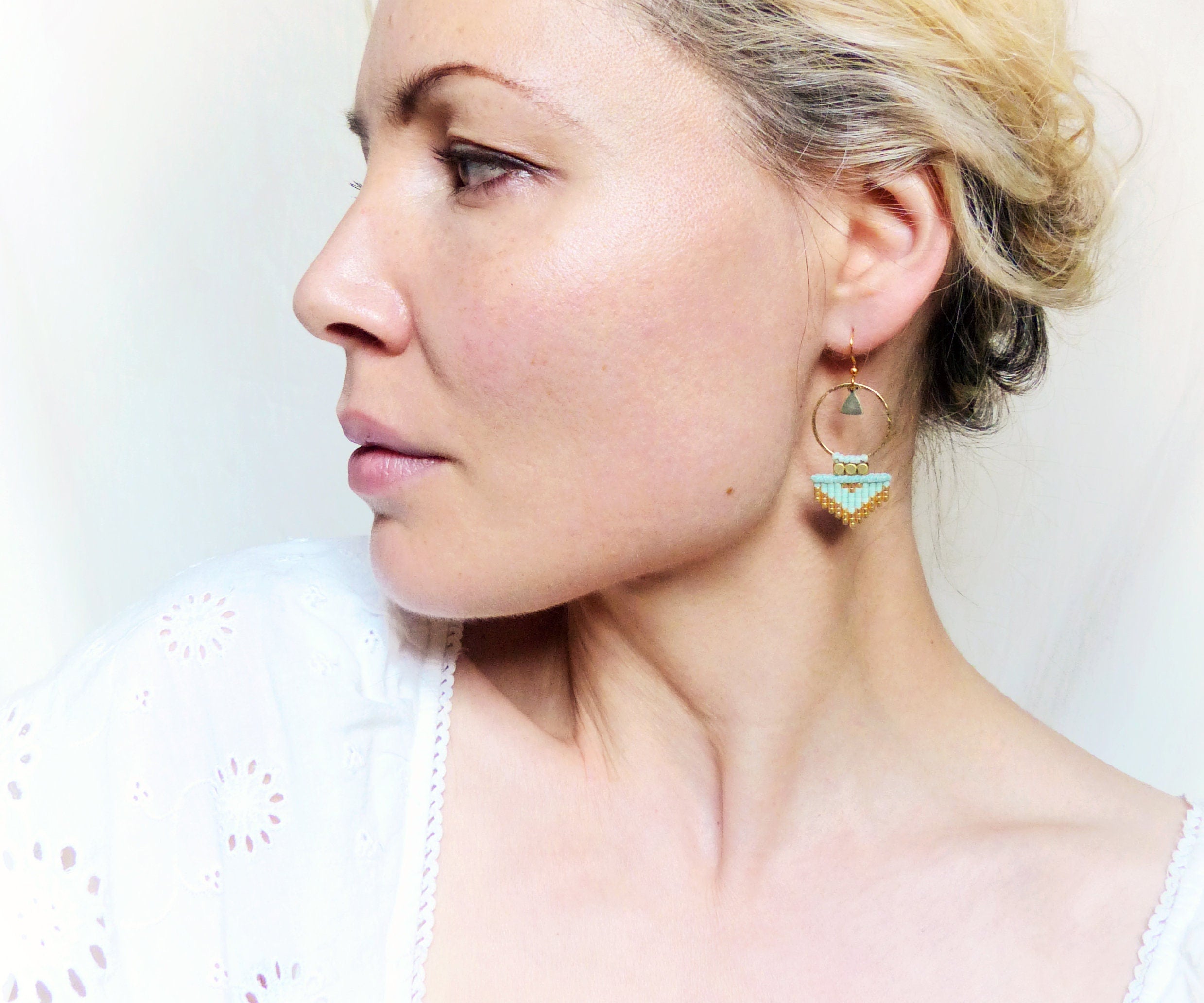 Peut inclure: Une paire de boucles d'oreilles créoles dorées avec un pendentif géométrique turquoise et or. Le pendentif est de forme triangulaire avec un plus petit triangle en haut. Les boucles d'oreilles sont suspendues à l'oreille d'une personne.
