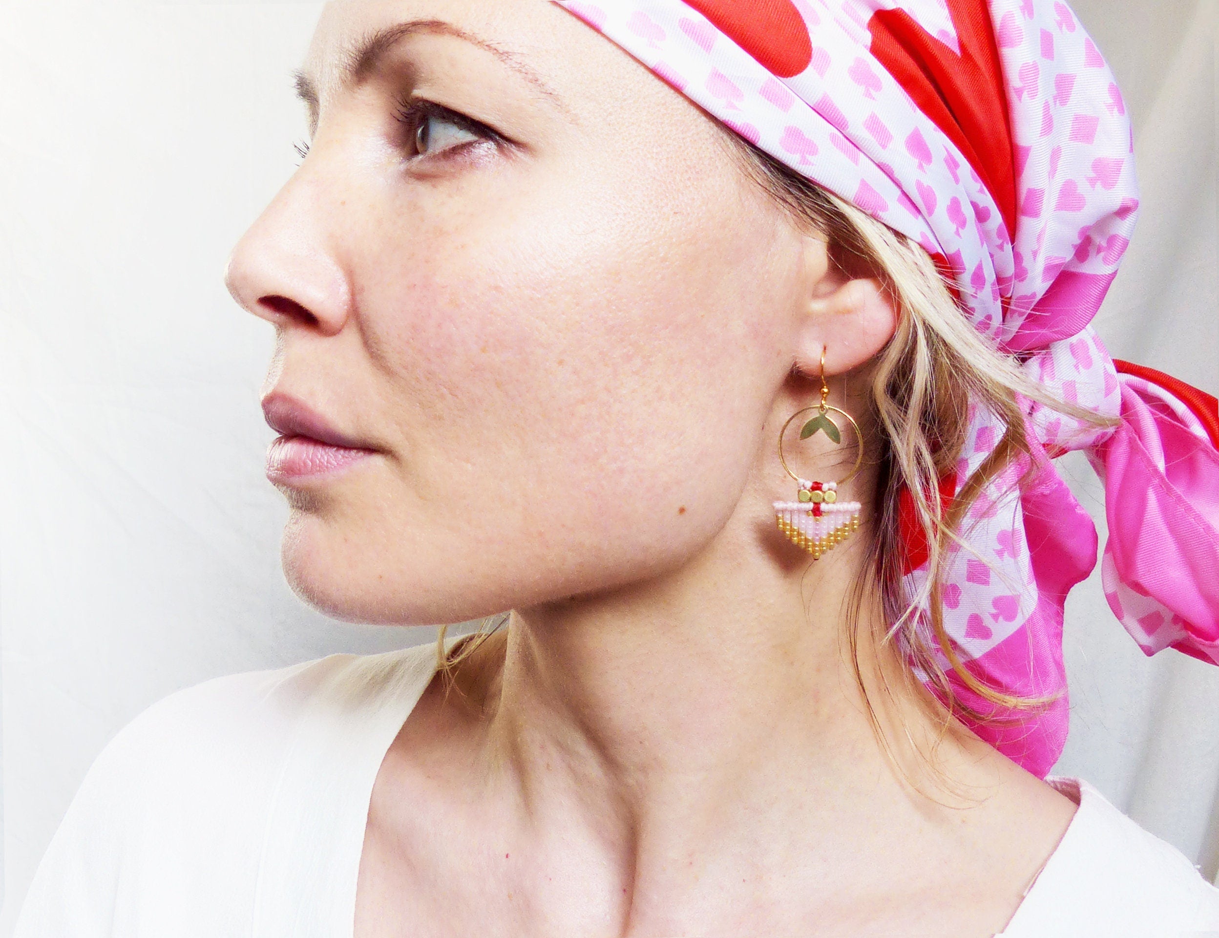 Peut inclure: Une femme portant un foulard à motifs roses et rouges avec des boucles d'oreilles en perles dorées et roses. Les boucles d'oreilles présentent un petit cercle avec une forme de cœur et un pendentif triangulaire avec une frange.