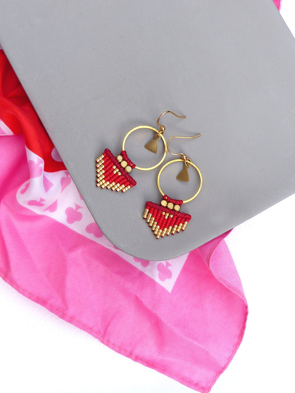 Peut inclure: Une paire de boucles d'oreilles en perles rouges et dorées avec un motif géométrique. Les boucles d'oreilles sont suspendues à des cercles dorés.
