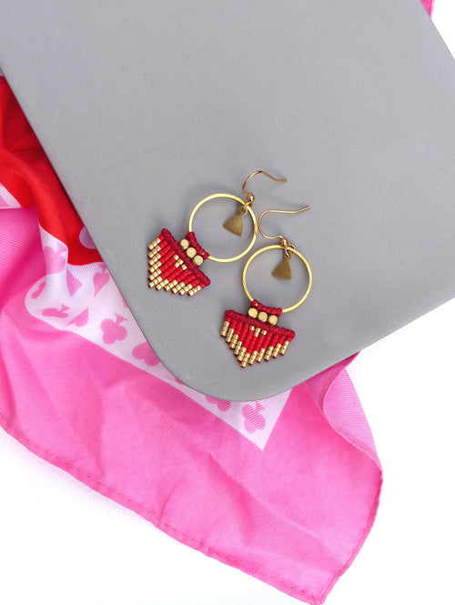 Peut inclure: Une paire de boucles d'oreilles en perles rouges et dorées avec un motif géométrique. Les boucles d'oreilles sont suspendues à des cercles dorés.