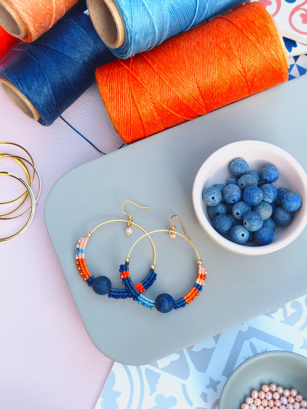 Peut inclure: Une paire de boucles d'oreilles créoles dorées avec des perles bleues, orange et roses. Les boucles d'oreilles sont sur une surface grise avec un bol blanc de perles bleues et un bol blanc de perles roses en arrière-plan.