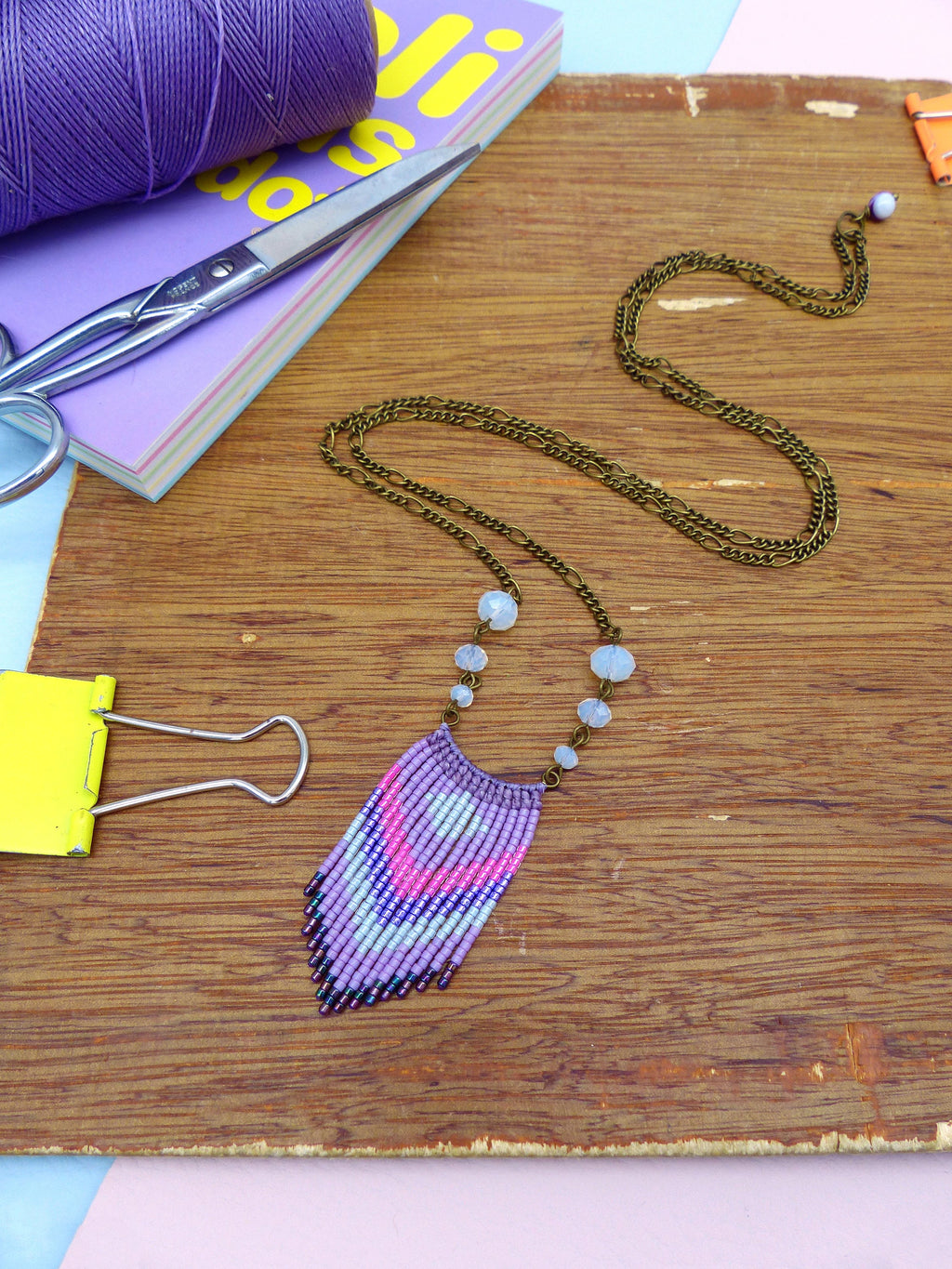 Peut inclure: Un collier à franges en perles violettes et blanches avec une chaîne en laiton et des perles de verre bleu clair. Le collier est sur une surface en bois.