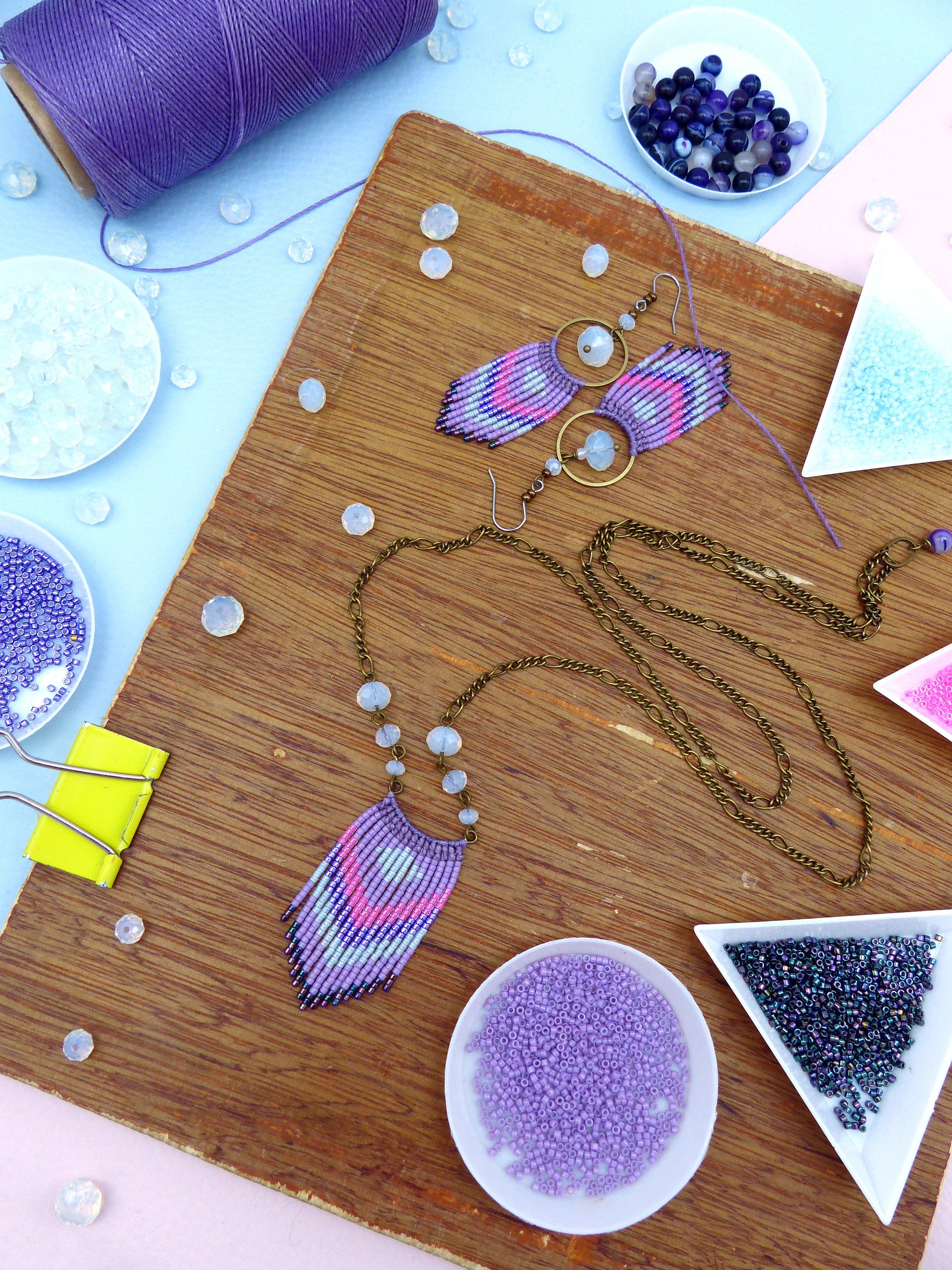 Peut inclure: Un plateau en bois avec une variété de perles et de fournitures pour la fabrication de bijoux. Il y a plusieurs petits bols remplis de perles de différentes couleurs, notamment du violet, du bleu et du noir. Il y a aussi un collier et une paire de boucles d'oreilles en perles violettes et bleues.