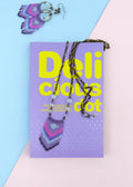 Peut inclure: Un collier de perles bleu et violet avec une chaîne argentée et deux perles de verre bleu clair. Le collier est posé sur un livre violet avec le texte "Delicious dot" en lettres jaunes.