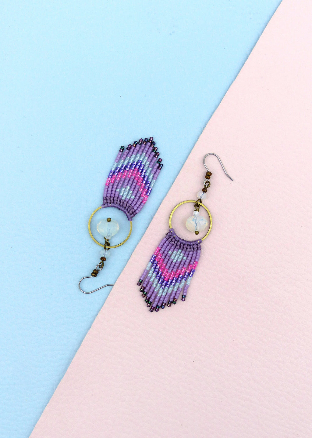 Peut inclure: Une paire de boucles d'oreilles en perles violettes, roses et blanches avec un charme en cristal transparent et un anneau en or. Les boucles d'oreilles sont sur un fond bleu clair et rose.