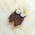 Peut inclure: Une paire de boucles d'oreilles créoles dorées avec un pendentif noir en perles brunes et dorées irisées. Chaque boucle d'oreille est ornée d'un petit charme en perle blanche.