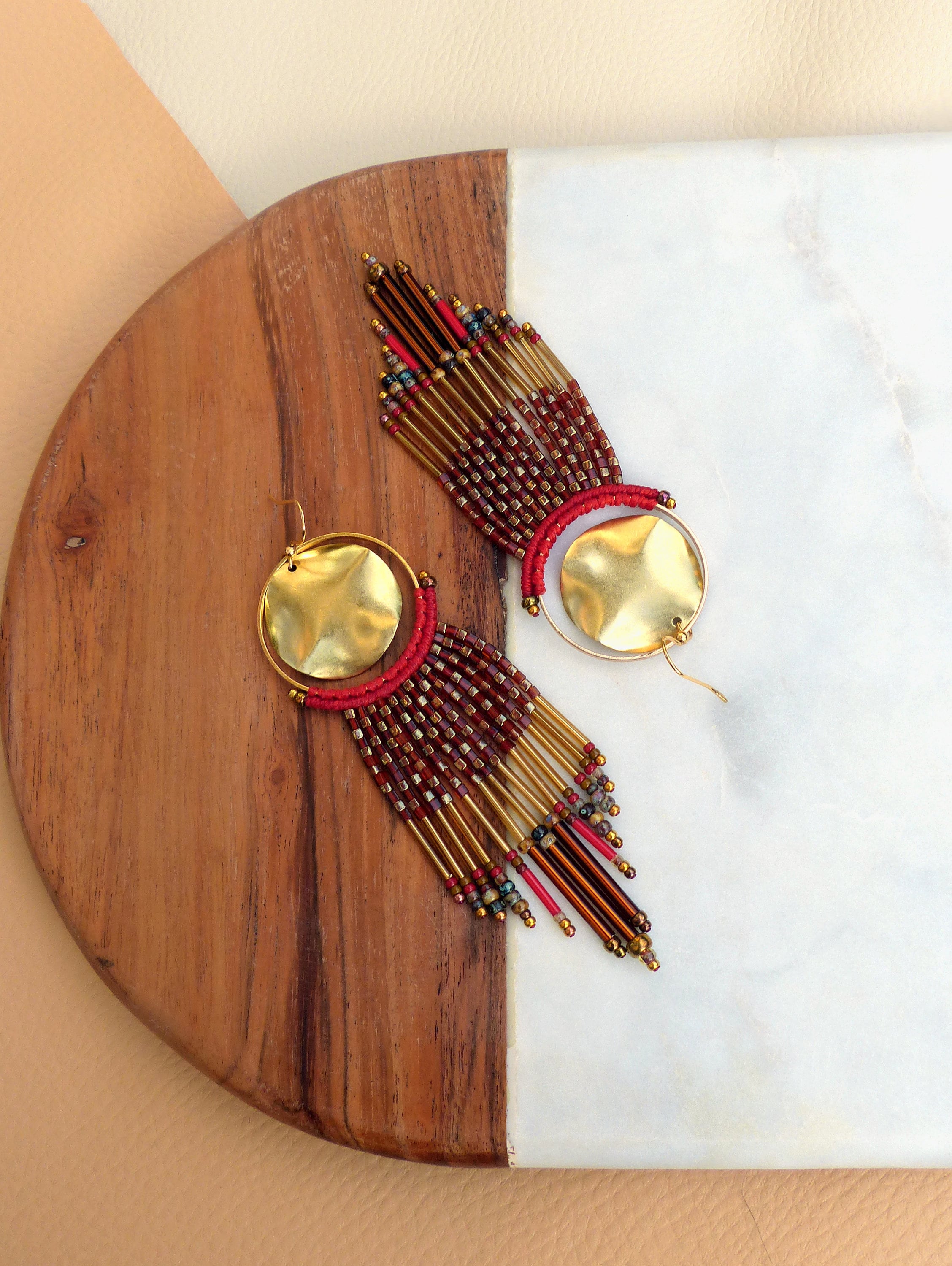 Peut inclure: Une paire de longues boucles d'oreilles en perles avec des accents dorés. Les boucles d'oreilles présentent un grand disque rond en or avec une texture martelée et une frange de perles rouges, dorées et brunes.