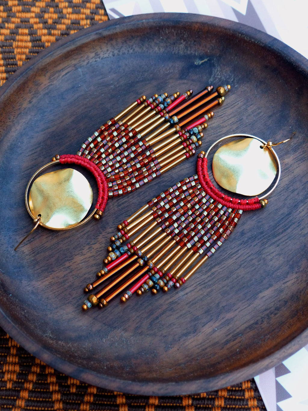 Peut inclure: Une paire de longues boucles d'oreilles en perles avec des anneaux dorés et des perles rouges et dorées. Les boucles d'oreilles sont suspendues à une surface en bois.