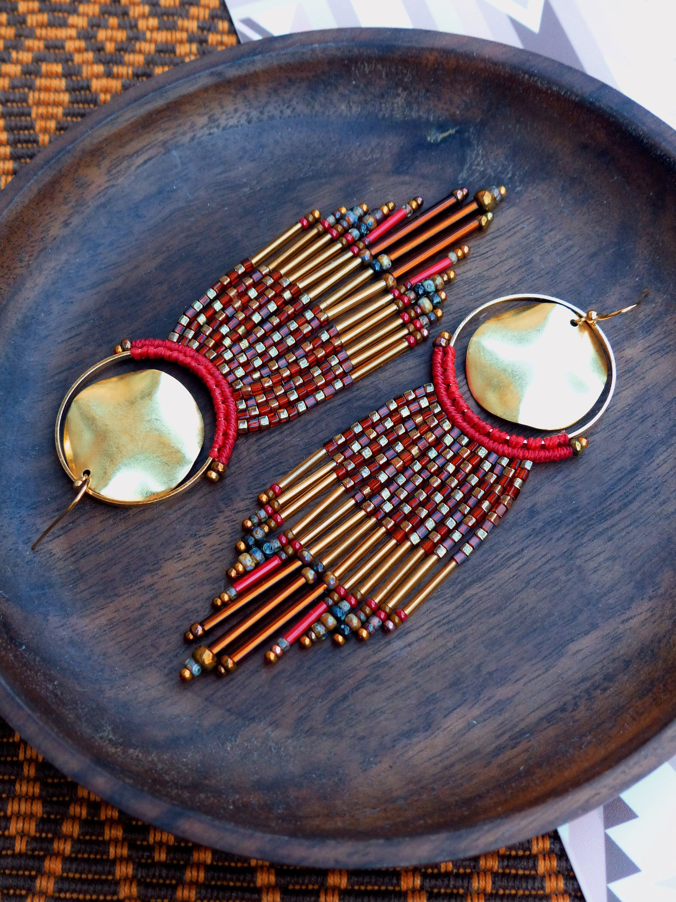 Peut inclure: Une paire de longues boucles d'oreilles en perles avec des anneaux dorés et des perles rouges et dorées. Les boucles d'oreilles sont suspendues à une surface en bois.