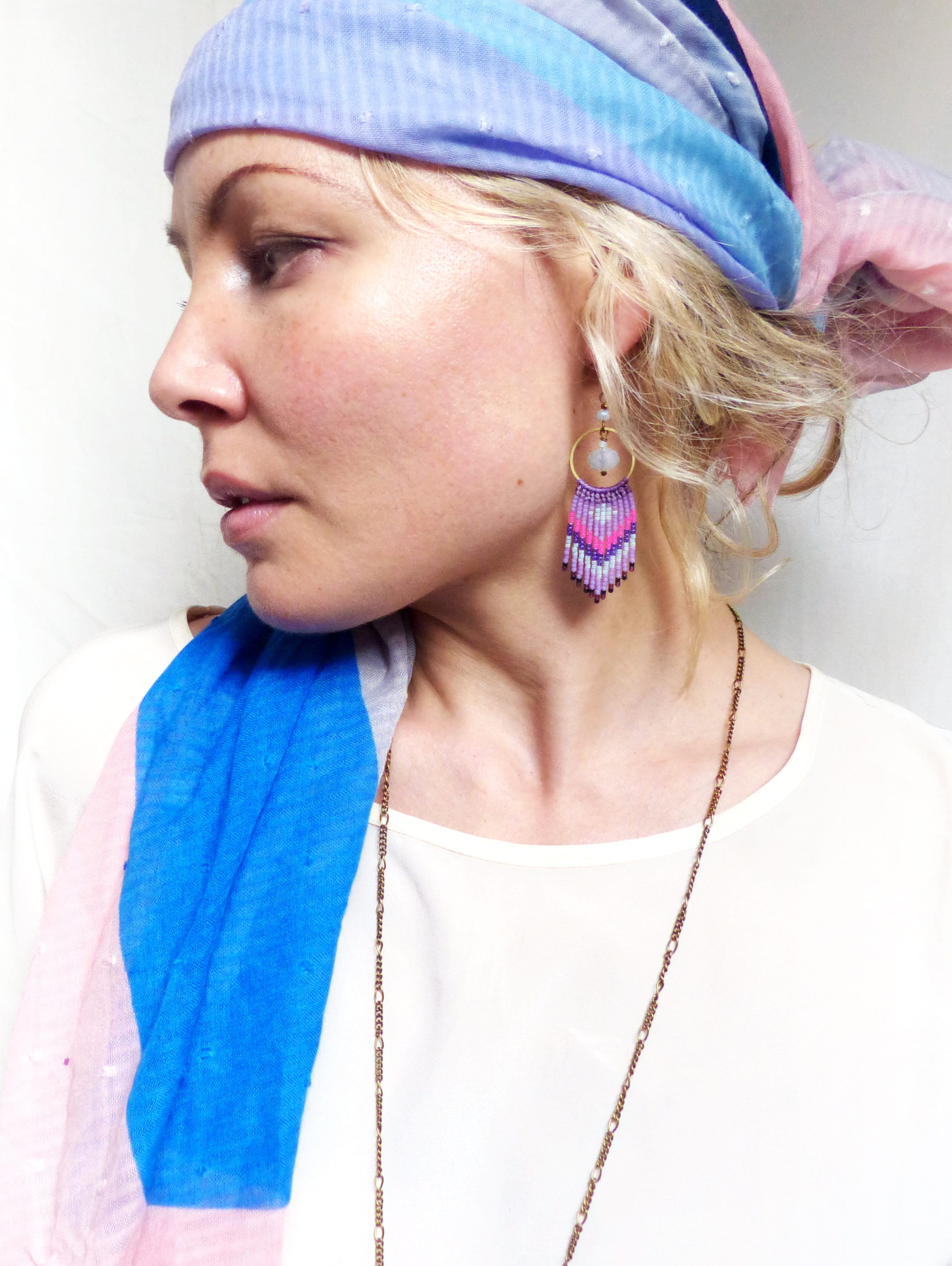 Peut inclure: Une femme portant une écharpe bleue et rose avec une chemise blanche et un collier de chaîne en or. Elle porte des boucles d'oreilles en perles violettes et roses avec une pierre blanche.