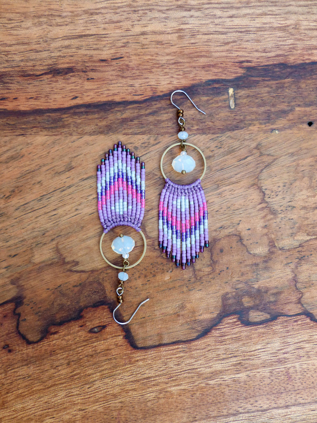 Peut inclure: Une paire de boucles d'oreilles en perles violettes et blanches avec un anneau en or et un pendentif en pierre transparente. Les boucles d'oreilles sont suspendues à une surface en bois.