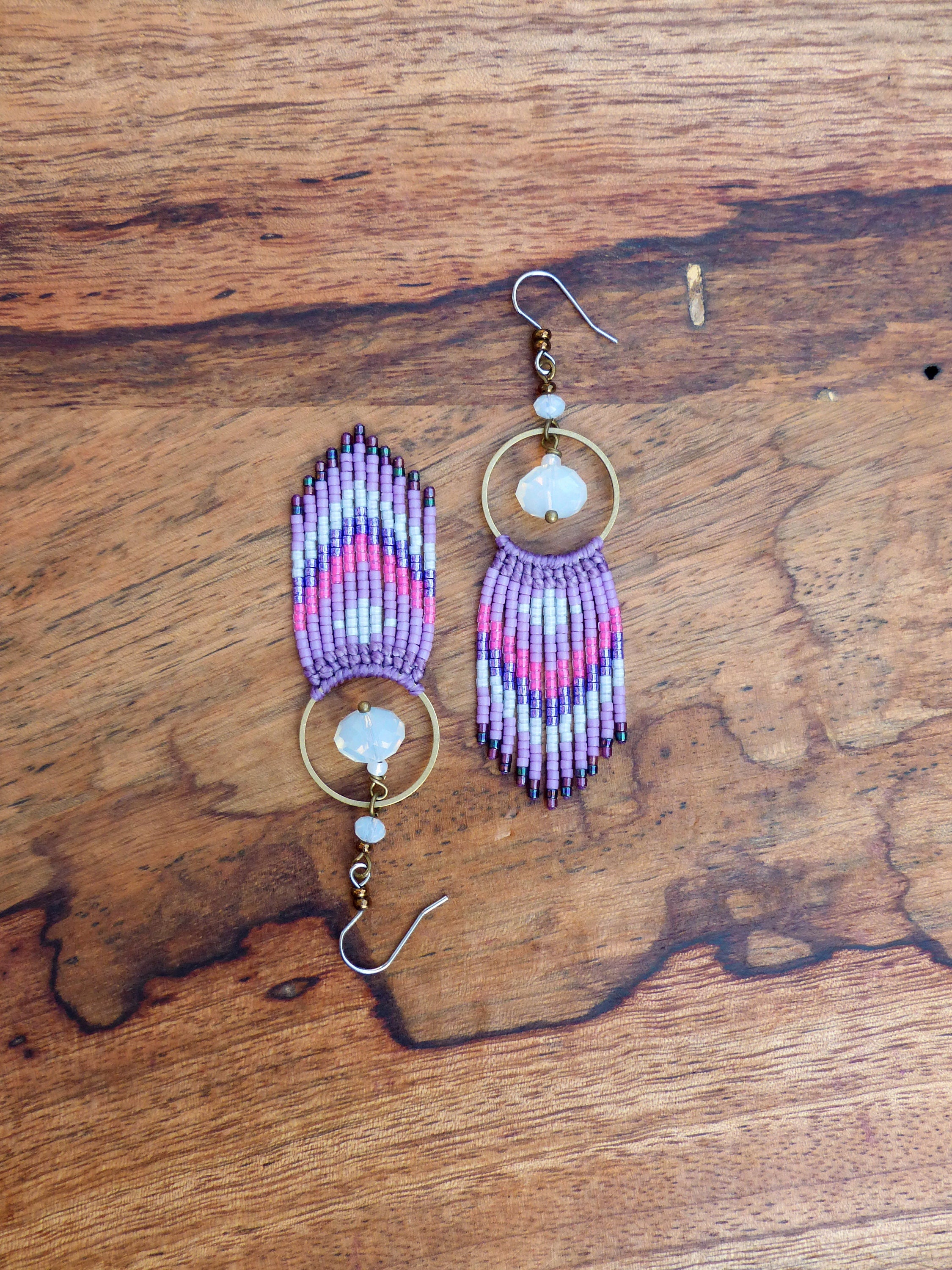 Peut inclure: Une paire de boucles d'oreilles en perles violettes et blanches avec un anneau en or et un pendentif en pierre transparente. Les boucles d'oreilles sont suspendues à une surface en bois.