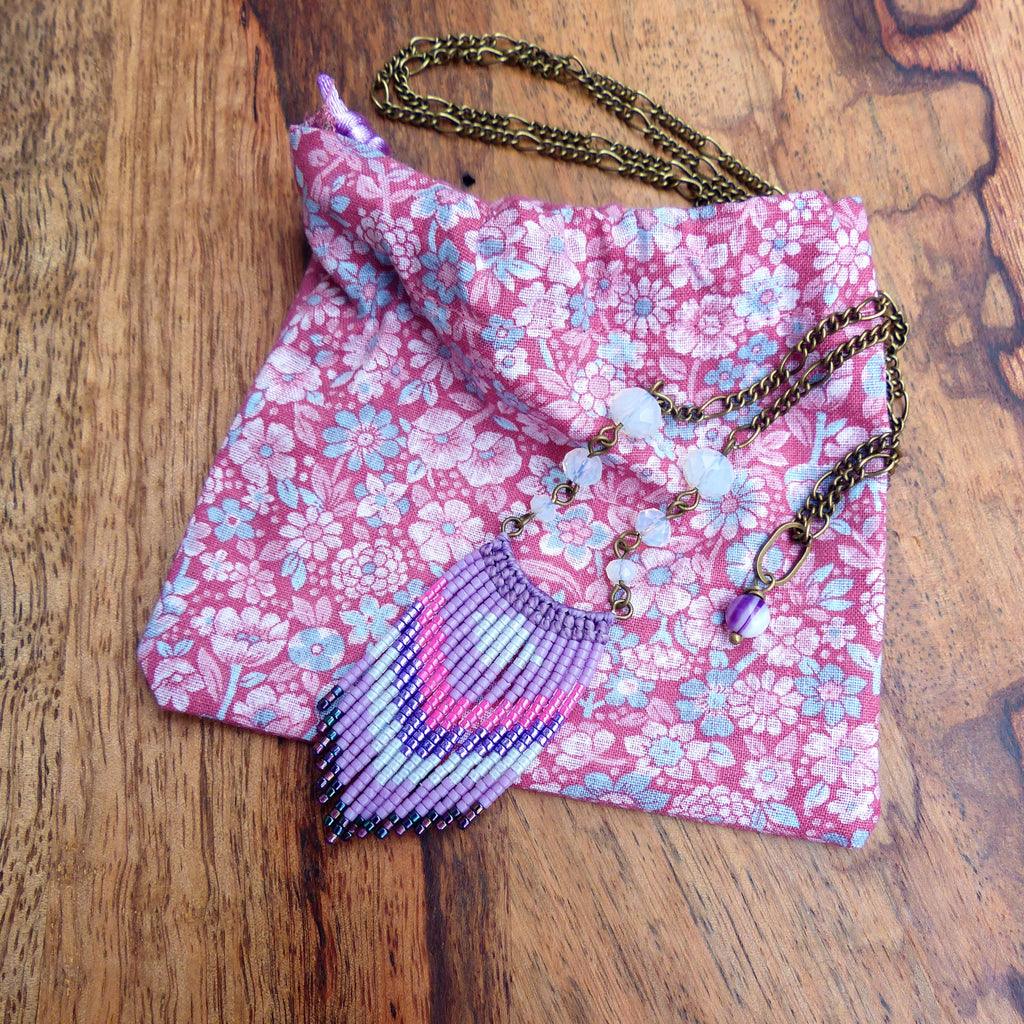 Peut inclure: Un collier à franges de perles violettes et roses avec un pendentif en pierre blanche et une chaîne en laiton. Le collier est présenté sur une pochette en tissu floral rose.