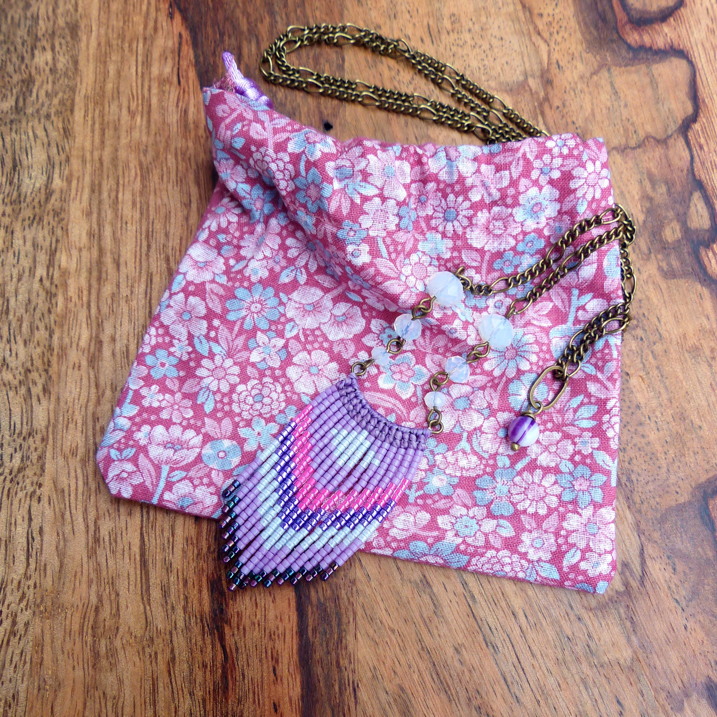 Peut inclure: Un collier à franges de perles violettes et roses avec un pendentif en pierre blanche et une chaîne en laiton. Le collier est présenté sur une pochette en tissu floral rose.