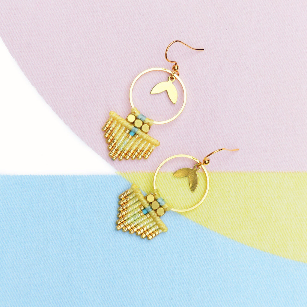 Peut inclure: Une paire de boucles d'oreilles faites à la main avec un design géométrique. Les boucles d'oreilles comportent un cercle doré avec un charme en forme de feuille. La partie inférieure est un motif tissé en perles jaunes, dorées et bleu clair. Le fond est un bloc de couleurs pastel.