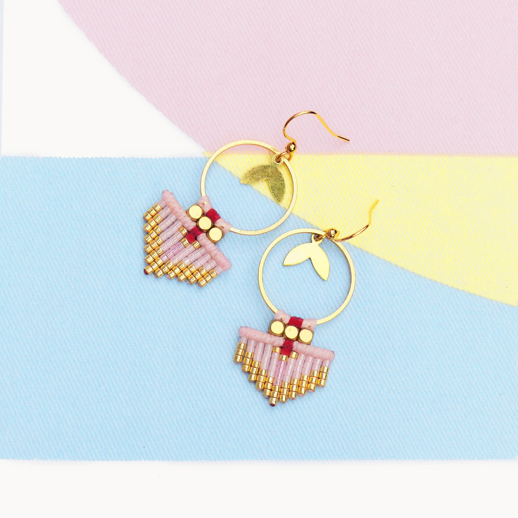 Peut inclure: Une paire de boucles d'oreilles faites à la main avec des cercles dorés et des détails de feuilles. Chaque boucle d'oreille présente une forme d'éventail perlé rose avec des accents dorés et rouges. Le fond est un bloc de couleurs pastel bleu, rose et jaune.
