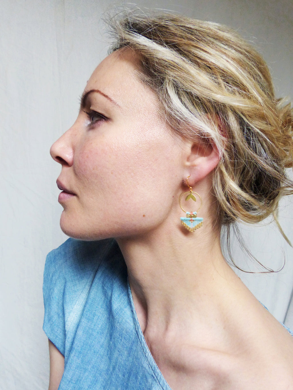 Peut inclure: Boucles d'oreilles dorées avec un motif géométrique. Les boucles d'oreilles présentent une section perlée bleu clair, une feuille dorée et de petites perles rouges. La personne porte un haut bleu.