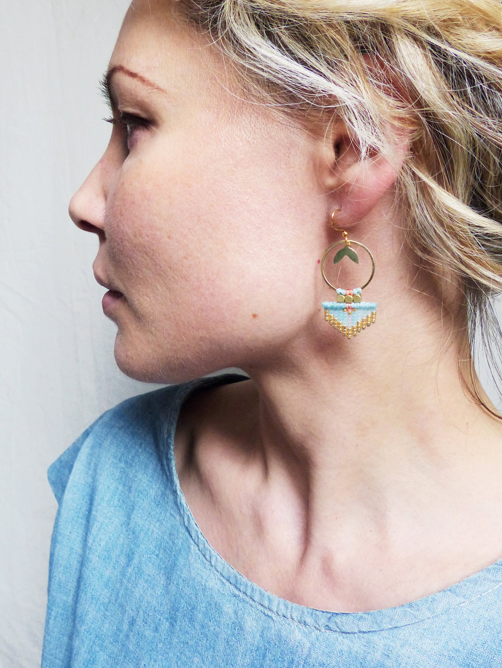 Peut inclure: Boucles d'oreilles dorées avec un design géométrique. Les boucles d'oreilles présentent une section perlée bleu clair avec des accents dorés et roses, et un petit détail de feuille verte. Les boucles d'oreilles sont portées par une personne.