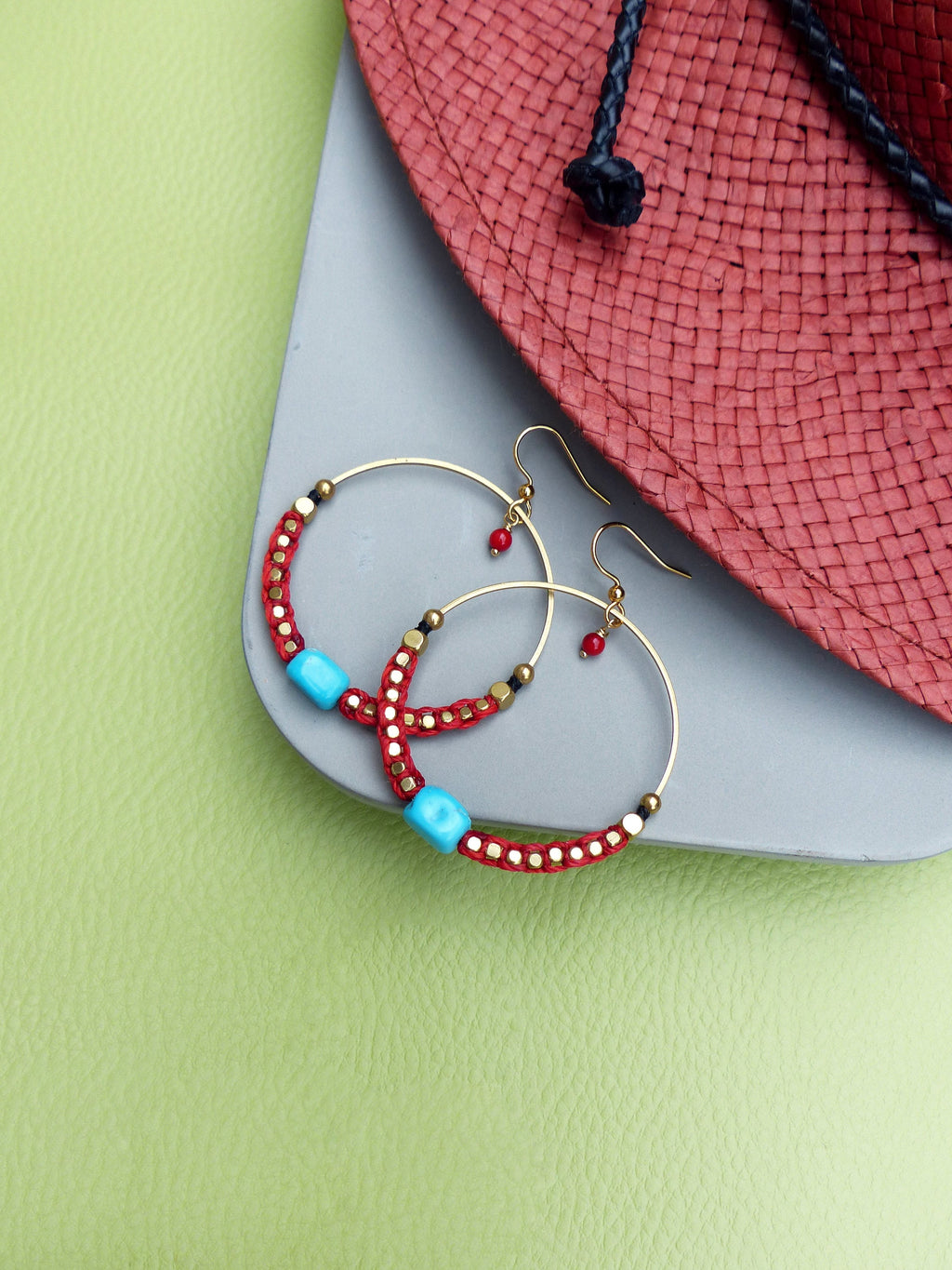 Peut inclure: Une paire de boucles d'oreilles créoles dorées avec des perles rouges et turquoise. Les boucles d'oreilles sont sur une surface grise avec un chapeau de paille rouge en arrière-plan.
