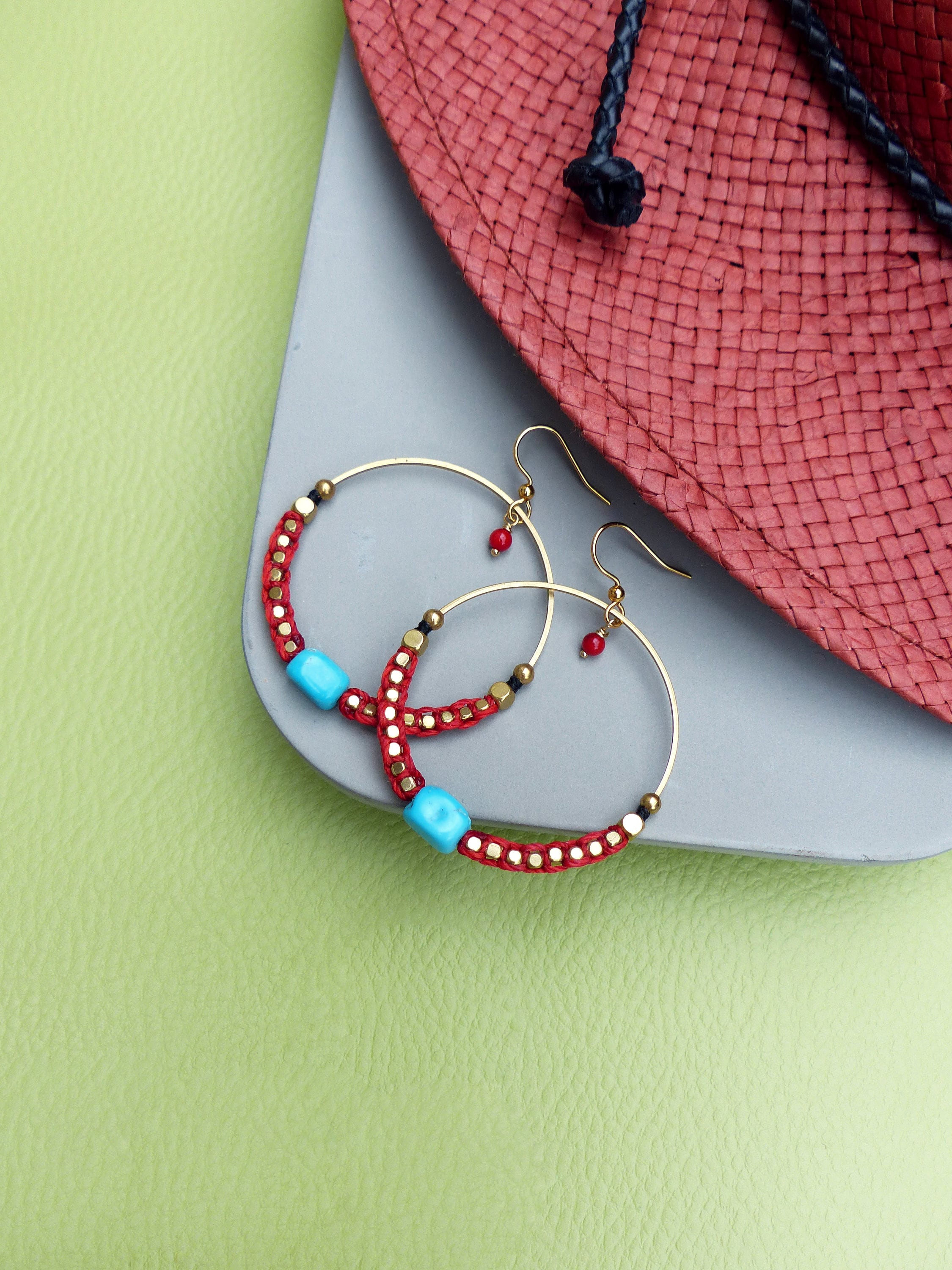 Peut inclure: Une paire de boucles d'oreilles créoles dorées avec des perles rouges et turquoise. Les boucles d'oreilles sont sur une surface grise avec un chapeau de paille rouge en arrière-plan.