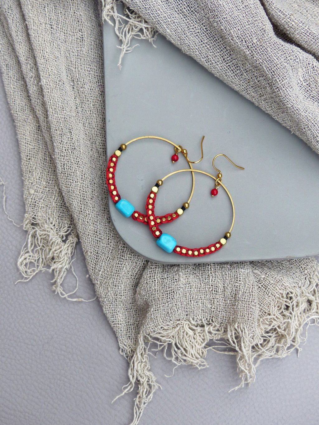 Peut inclure: Une paire de boucles d'oreilles créoles dorées avec des perles rouges et turquoise. Les boucles d'oreilles sont sur une surface grise.