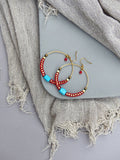 Peut inclure: Une paire de boucles d'oreilles créoles dorées avec des perles rouges et turquoise. Les boucles d'oreilles sont sur une surface grise.