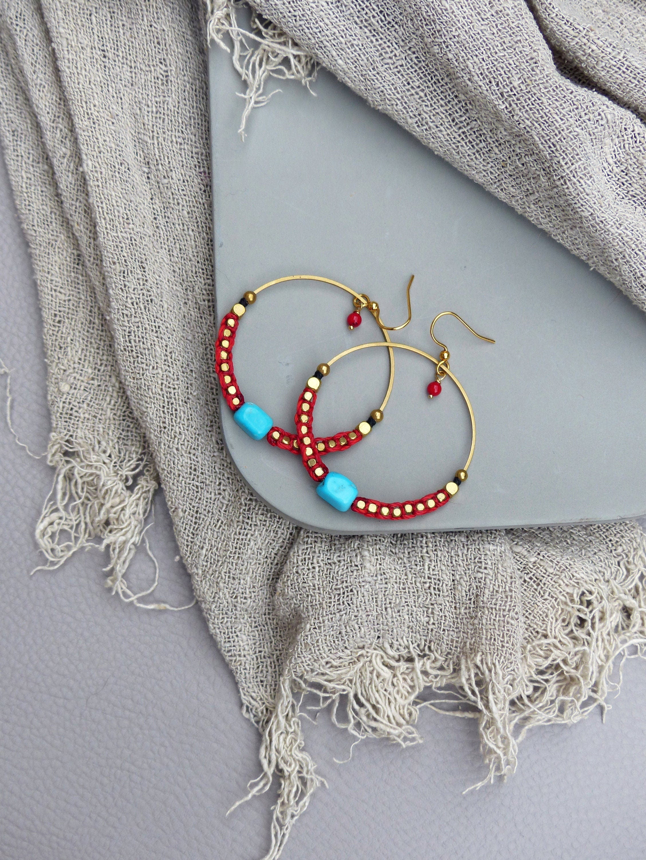 Peut inclure: Une paire de boucles d'oreilles créoles dorées avec des perles rouges et turquoise. Les boucles d'oreilles sont sur une surface grise.