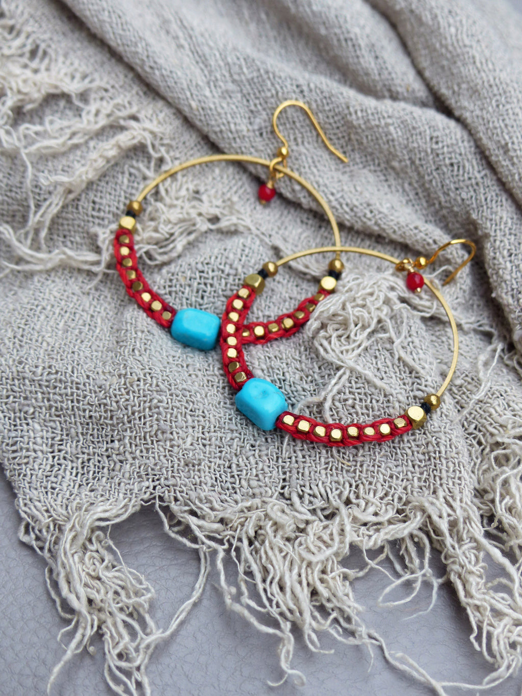 Peut inclure: Une paire de boucles d'oreilles créoles dorées avec des perles rouges et turquoise. Les boucles d'oreilles sont sur un fond de tissu gris clair.