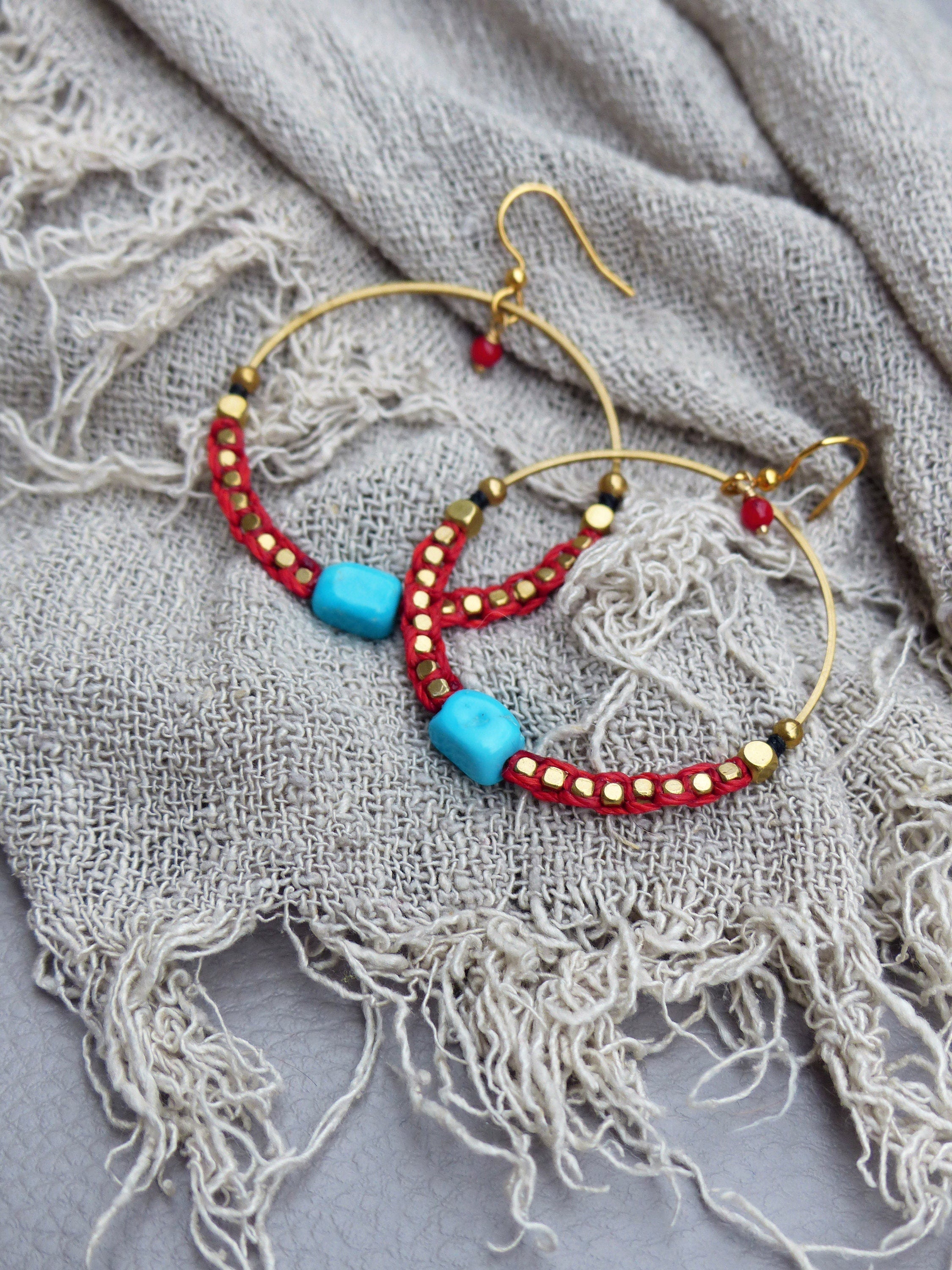 Peut inclure: Une paire de boucles d'oreilles créoles dorées avec des perles rouges et turquoise. Les boucles d'oreilles sont sur un fond de tissu gris clair.