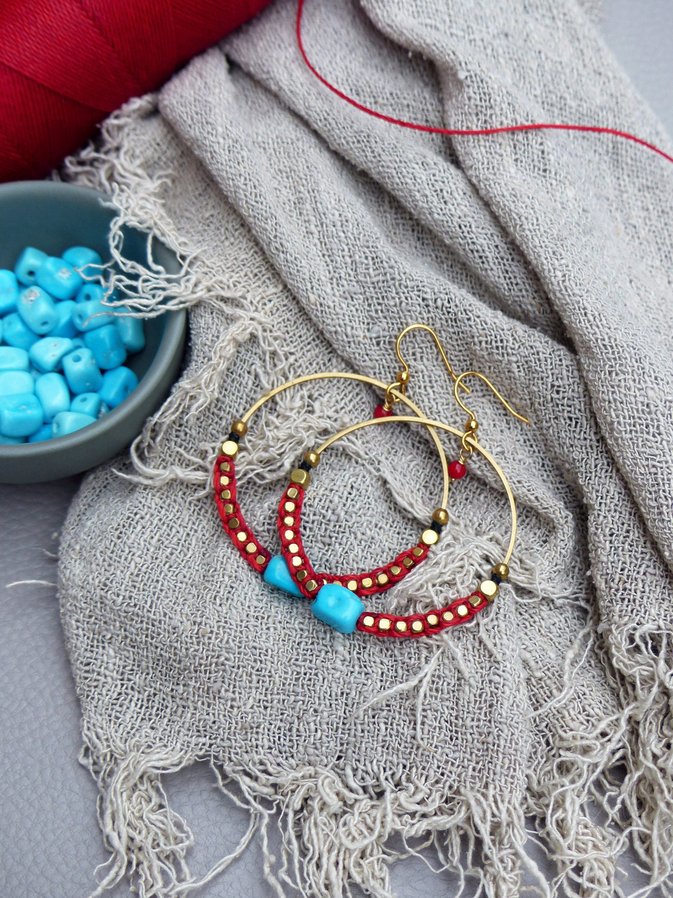 Peut inclure: Une paire de boucles d'oreilles créoles dorées avec des perles rouges et turquoise. Les boucles d'oreilles sont sur un fond de tissu gris clair.