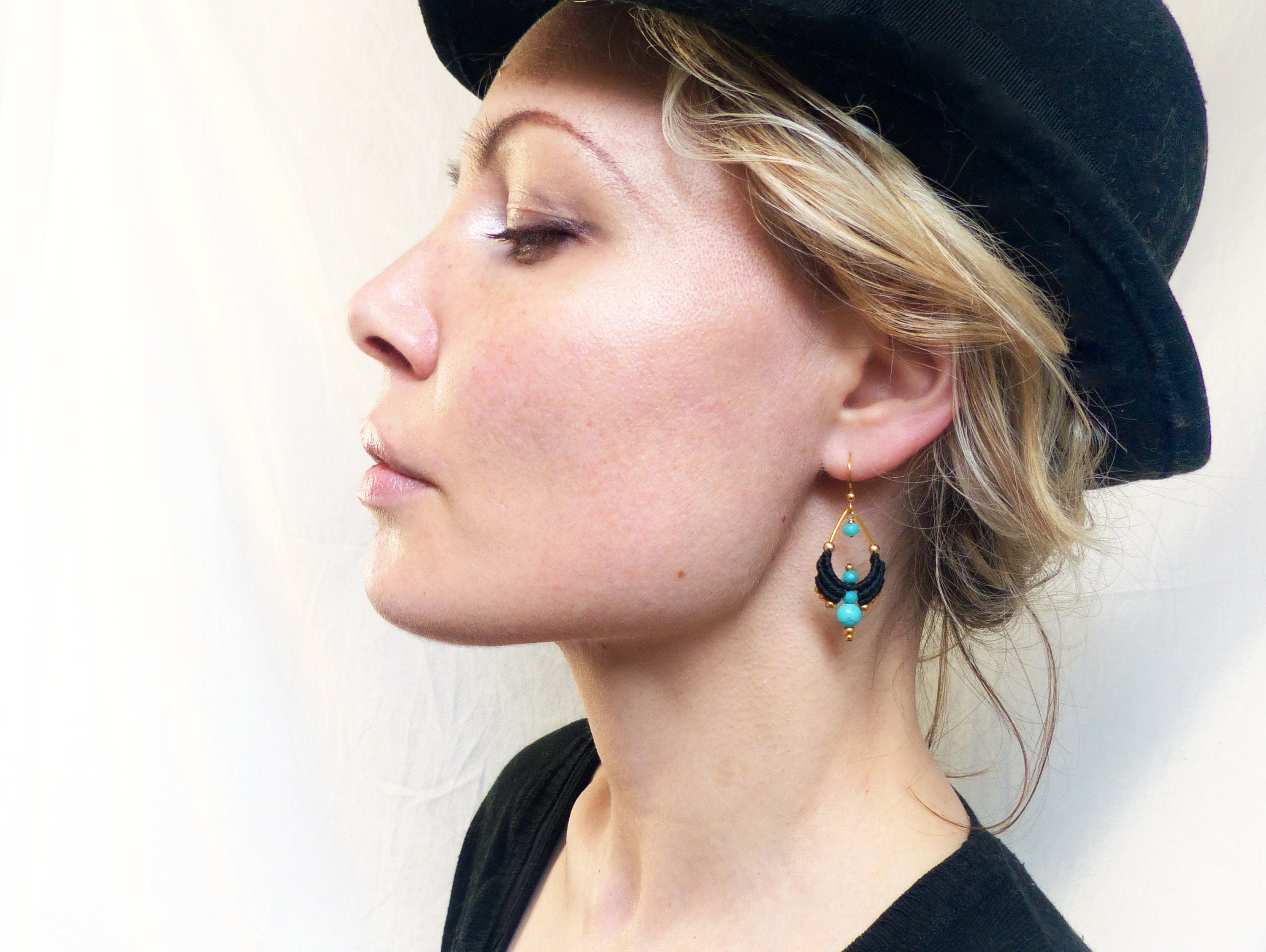 Peut inclure: Une femme portant un chapeau noir et un chemisier noir avec des boucles d'oreilles turquoise et dorées. Les boucles d'oreilles sont faites de fil noir et ont une forme de larme avec des perles turquoise.