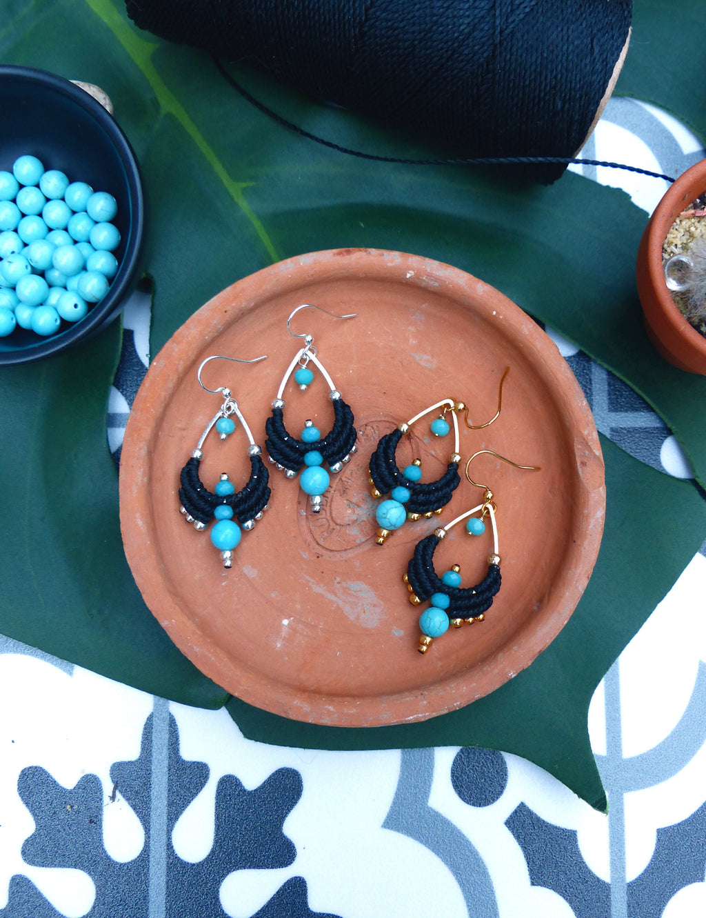Peut inclure: Une paire de boucles d'oreilles pendantes avec des perles turquoise et du macramé noir. Les boucles d'oreilles sont sur une assiette en terre cuite.