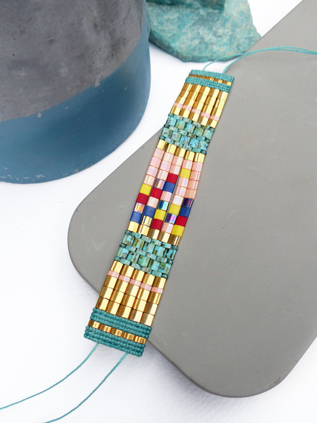 Peut inclure: Un bracelet de perles avec un motif rectangulaire. Il présente des perles dorées, roses, bleues, rouges et jaunes, enfilées sur un cordon turquoise. Le bracelet est présenté sur une surface grise. Le design est géométrique et coloré.