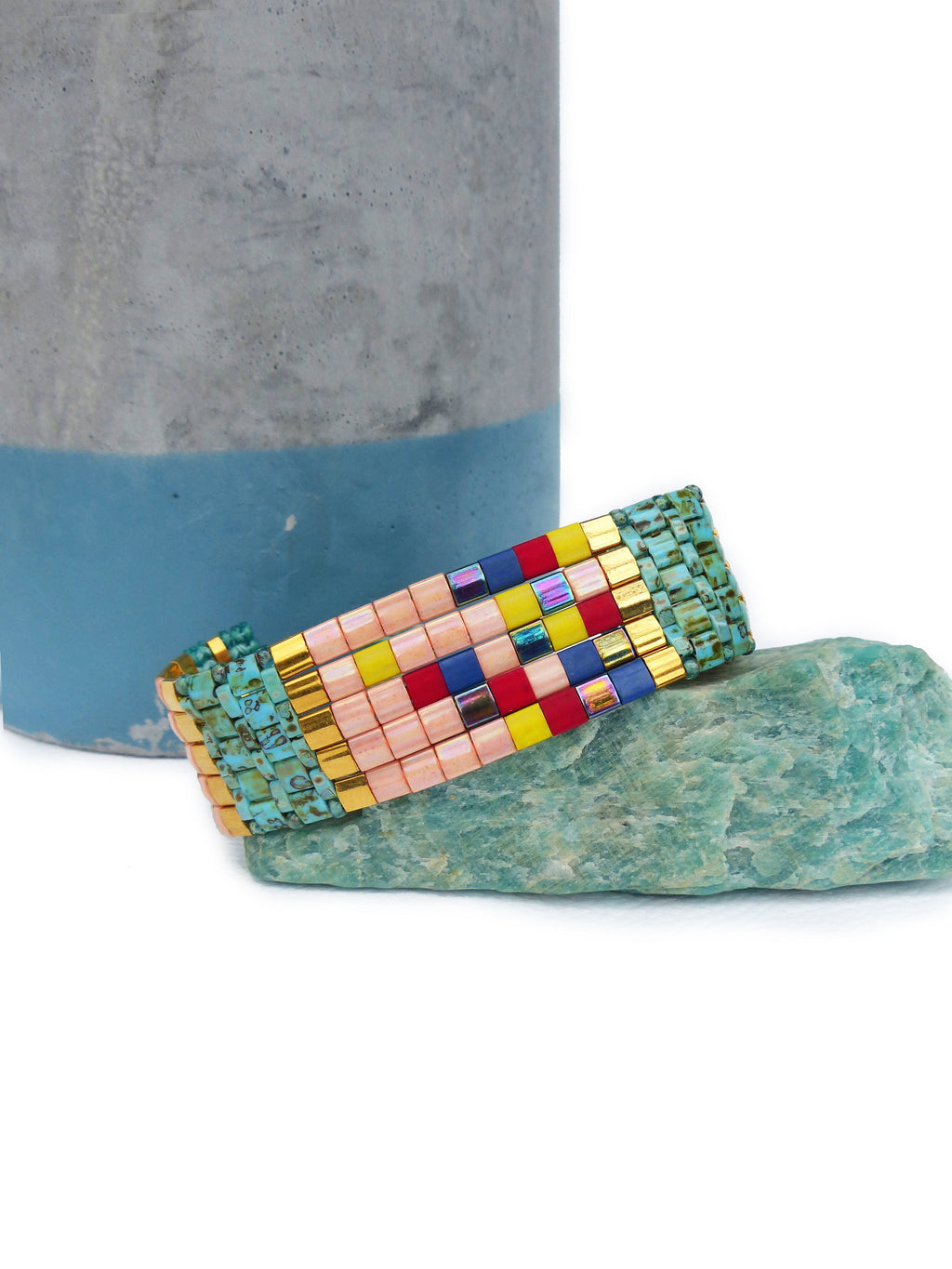 Peut inclure: Un bracelet de perles colorées avec un motif géométrique. Le bracelet présente un mélange de perles roses, jaunes, rouges, bleues et irisées, avec des accents dorés. Il est présenté sur une pierre vert clair.